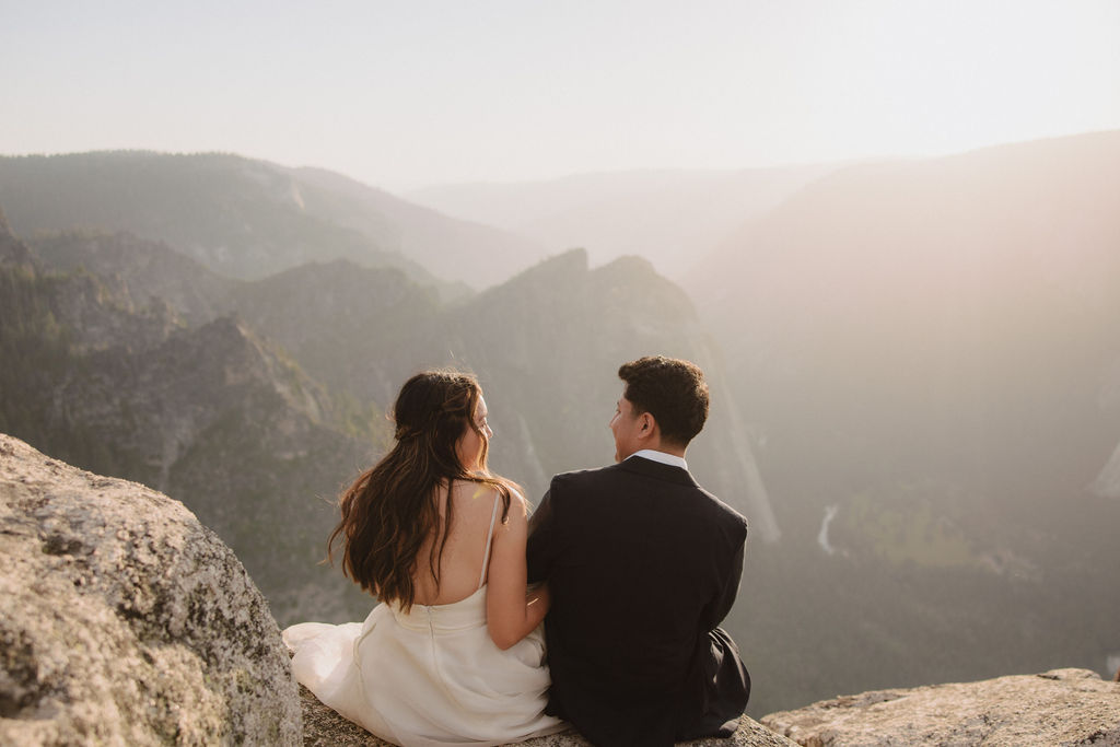 A couple in formal attire stands on the edge of a cliff, embracing and looking at each other, with vast mountainous scenery in the background under a hazy sky in Yosemite