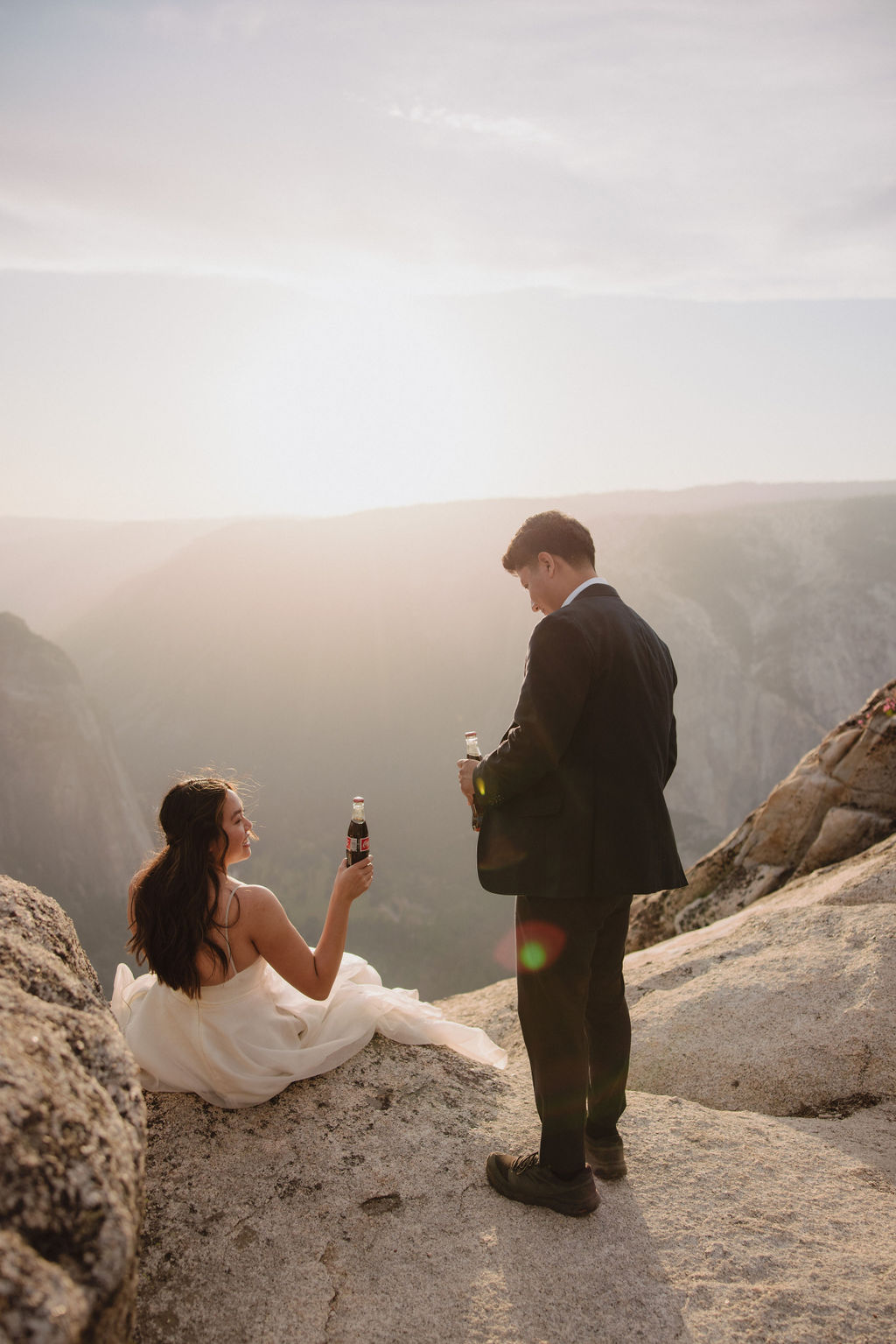 A couple in wedding attire stand on a rocky landscape. The woman in a white dress drinks from a bottle while the man in a black suit stands beside her holding a bottle.