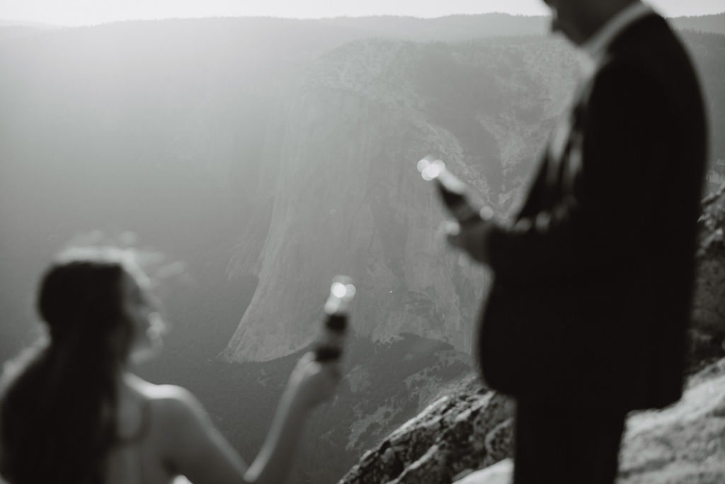 A bride and groom sit on a rocky cliff, dressed in wedding attire, sharing drinks from bottles with mountainous scenery in the background.