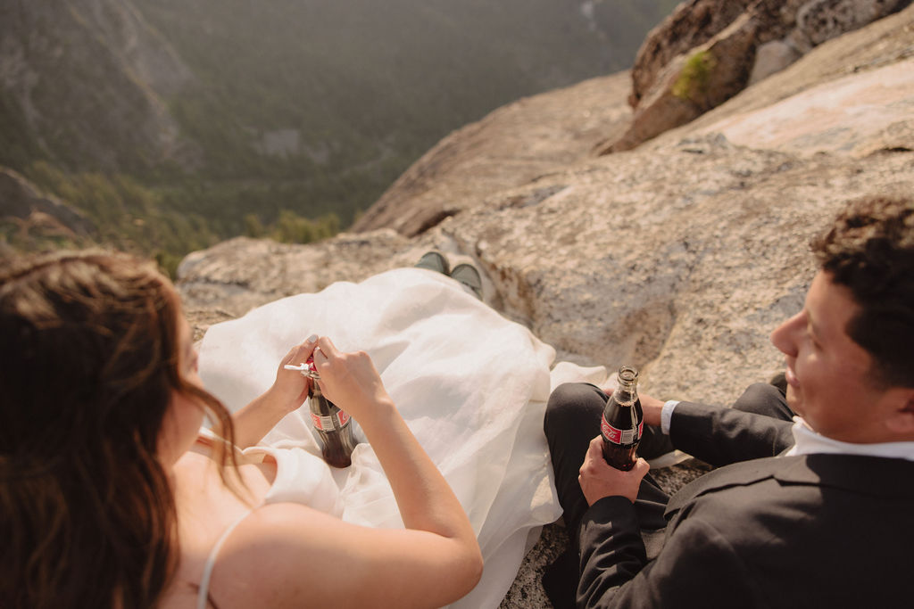 A bride and groom sit on a rocky cliff, dressed in wedding attire, sharing drinks from bottles with mountainous scenery in the background.