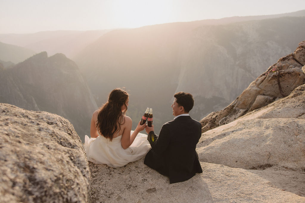 A couple in wedding attire stand on a rocky landscape. The woman in a white dress drinks from a bottle while the man in a black suit stands beside her holding a bottle.