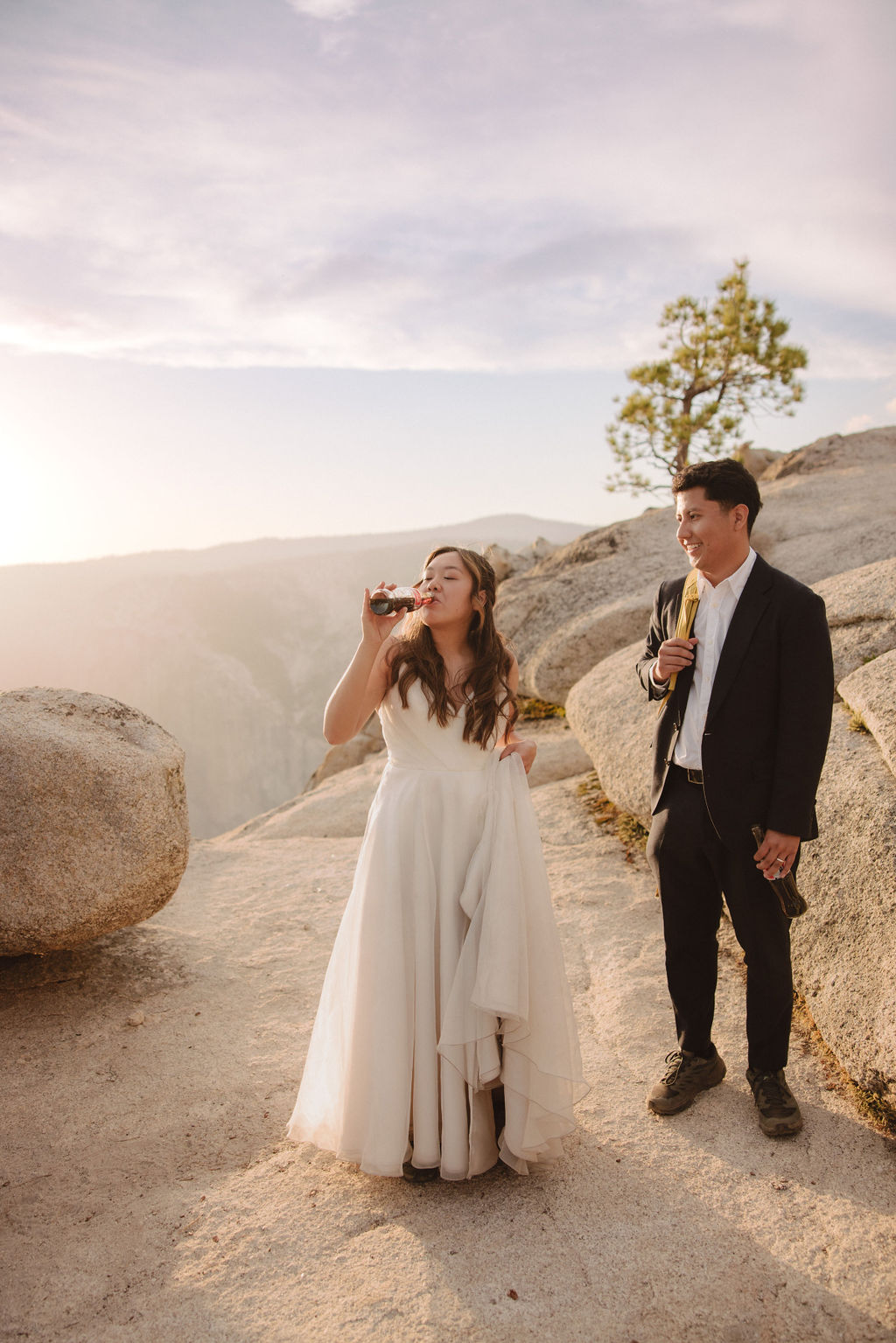 A couple in wedding attire stand on a rocky landscape. The woman in a white dress drinks from a bottle while the man in a black suit stands beside her holding a bottle.