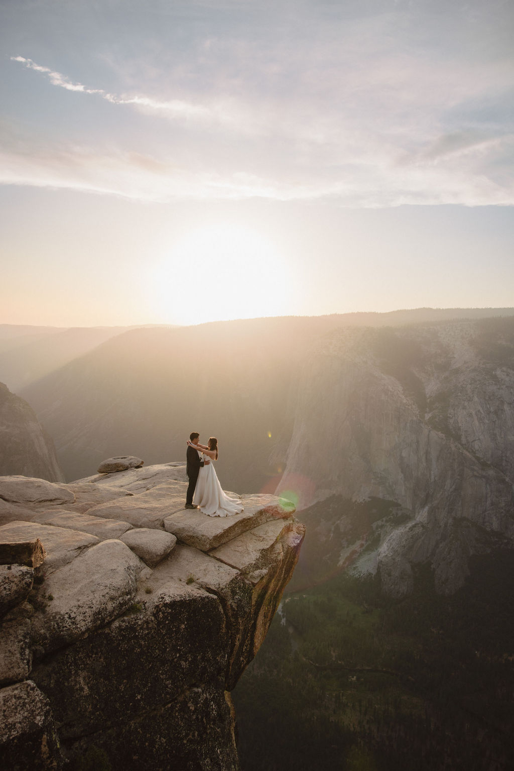 A bride and groom stand on a cliff edge, overlooking a vast landscape with mountains and a setting sun in the background for a Yosemite elopement