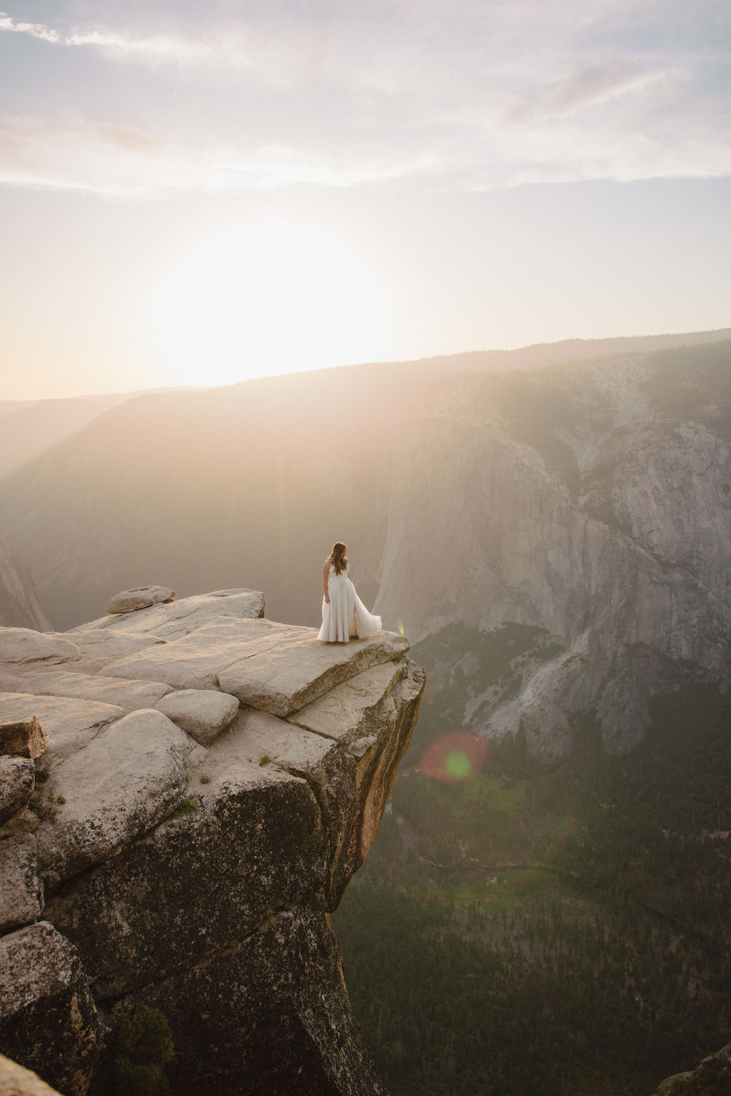 A bride and groom stand on a cliff edge, overlooking a vast landscape with mountains and a setting sun in the background for a Yosemite elopement