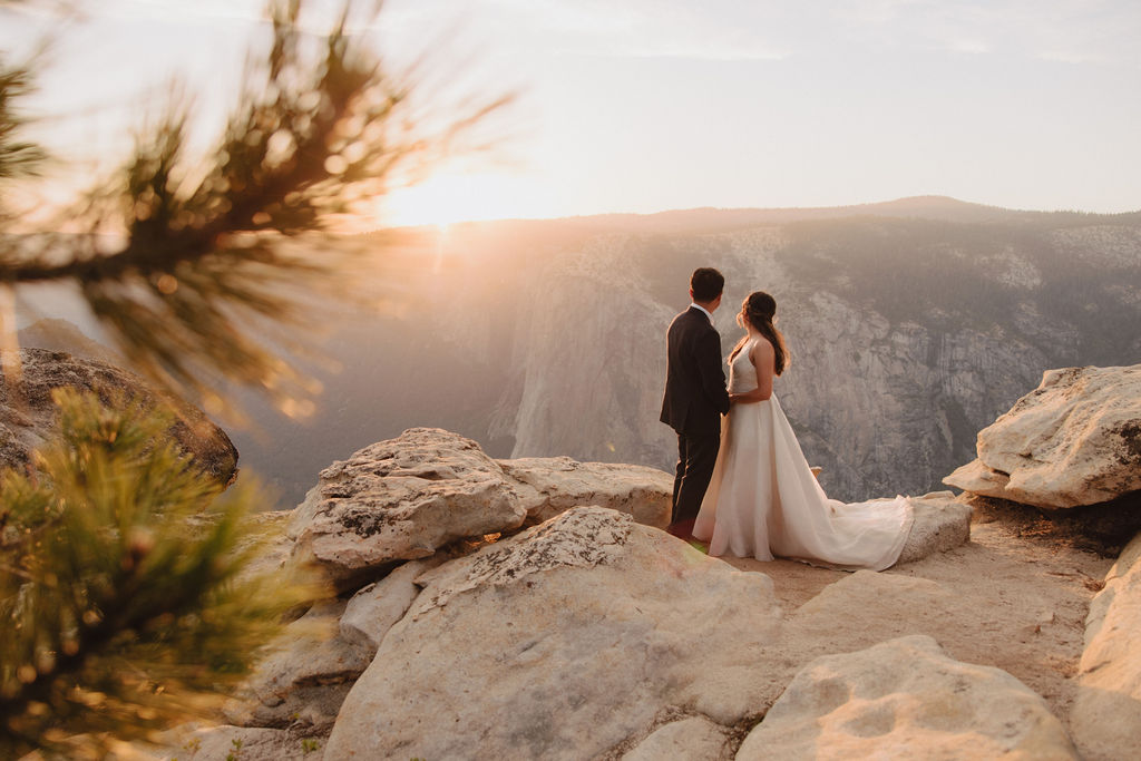 A bride and groom stand on a cliff edge, overlooking a vast landscape with mountains and a setting sun in the background for a Yosemite elopement