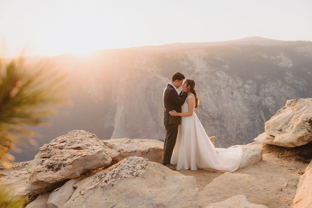 A couple in wedding attire embraces and kisses at sunset on a cliff with a mountain range in the background| tips for yosemite elopement