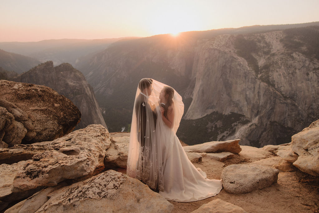 A bride and groom stand on a cliff edge, overlooking a vast landscape with mountains and a setting sun in the background for a Yosemite elopement