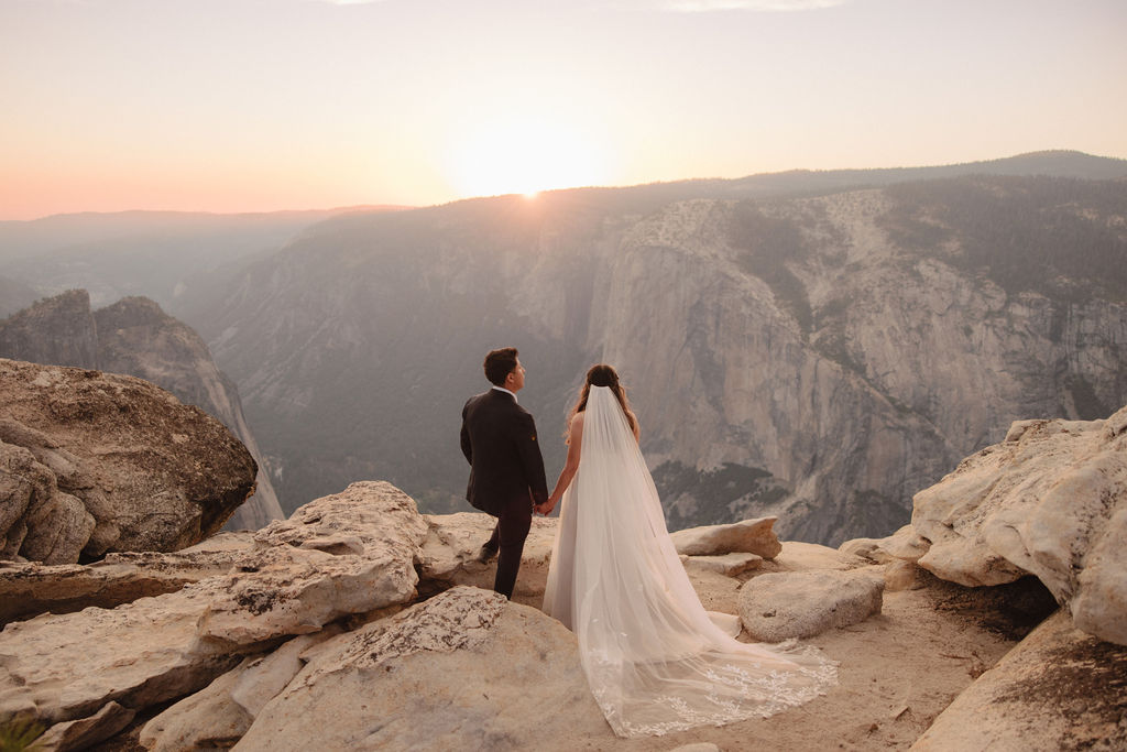 A bride and groom stand on a cliff edge, overlooking a vast landscape with mountains and a setting sun in the background for a Yosemite elopement