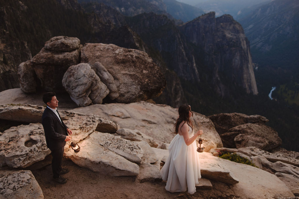 A couple in wedding attire stands on rocky terrain holding lanterns, overlooking a valley with cliffs and a distant river in Yosemite