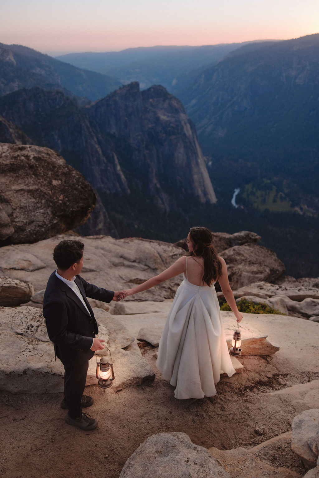 A couple in wedding attire stands on rocky terrain holding lanterns, overlooking a valley with cliffs and a distant river in Yosemite