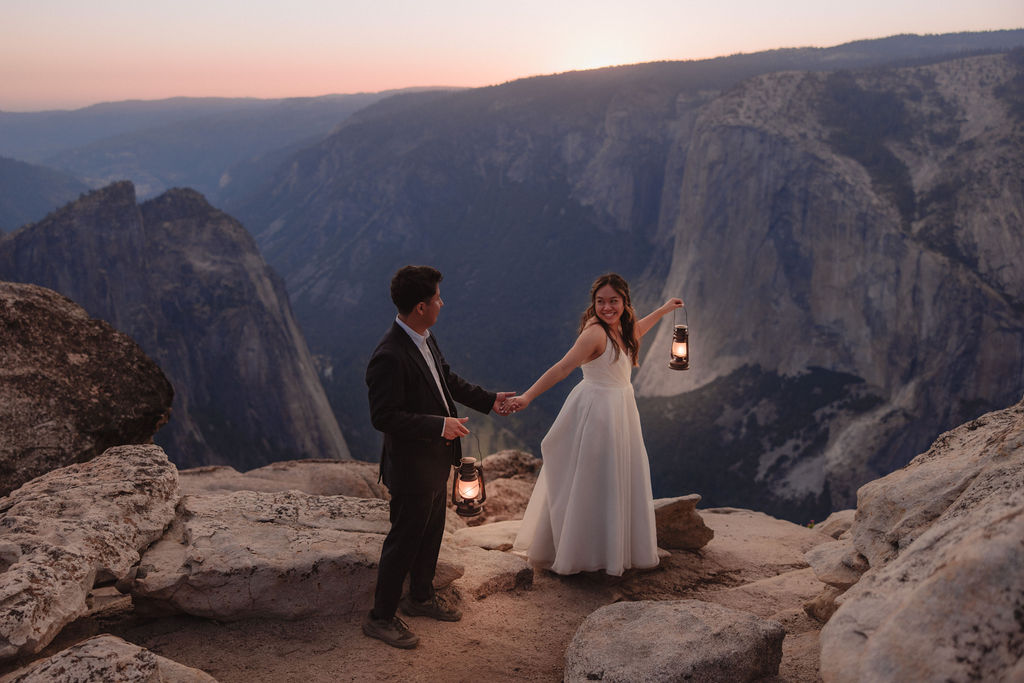 A couple in wedding attire stands on rocky terrain holding lanterns, overlooking a valley with cliffs and a distant river in Yosemite 