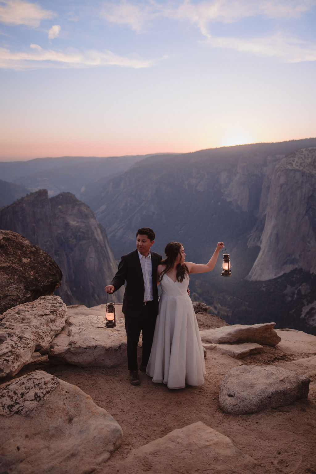 A couple in formal attire holds lanterns while standing on a rocky ledge overlooking a vast mountainous landscape at sunset.