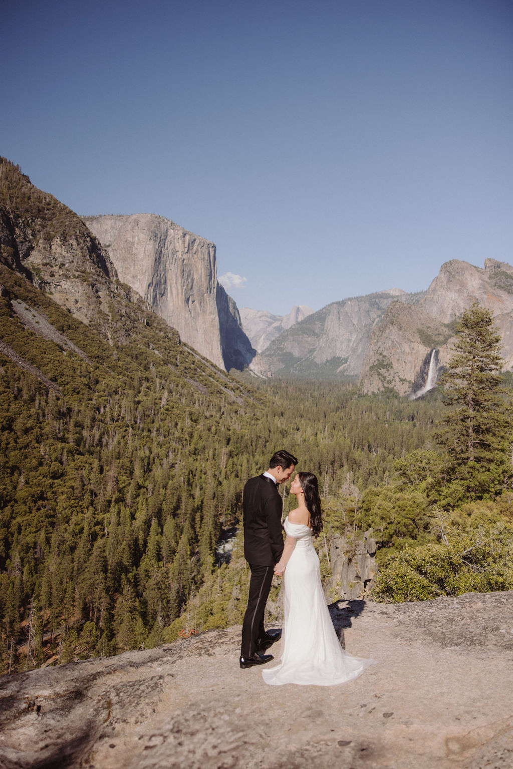 A bride and groom stand on a cliff edge in a mountainous landscape with snow-capped peaks and forests in the background. The bride wears a fur shawl over her gown, and the groom is in a dark suit at Tunnel view for their elopement at Yosemite national park 