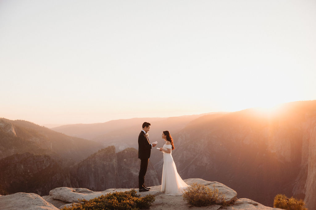 A bride and groom stand on a rocky cliff edge with mountains and a sunset in the background. | Tips for planning a stress free yosemite elopement 