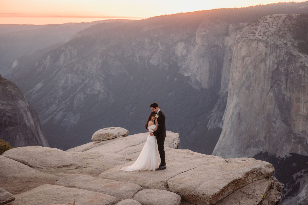 A bride and groom stand on a rocky cliff edge with mountains and a sunset in the background. | Tips for planning a stress free yosemite elopement 