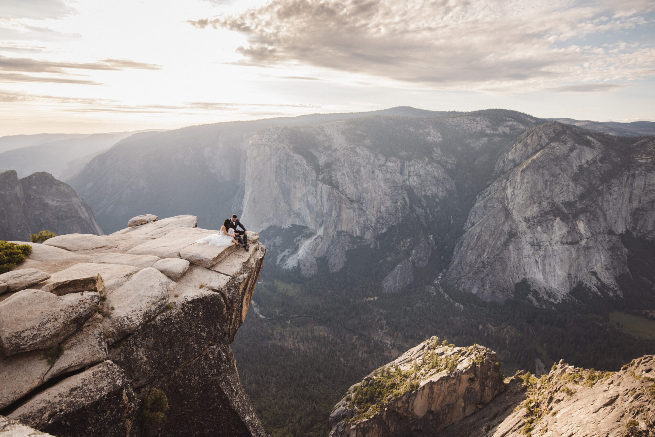 A bride and groom stand on a cliff edge overlooking a vast mountainous landscape at sunset at Taft point Yosemite national park 