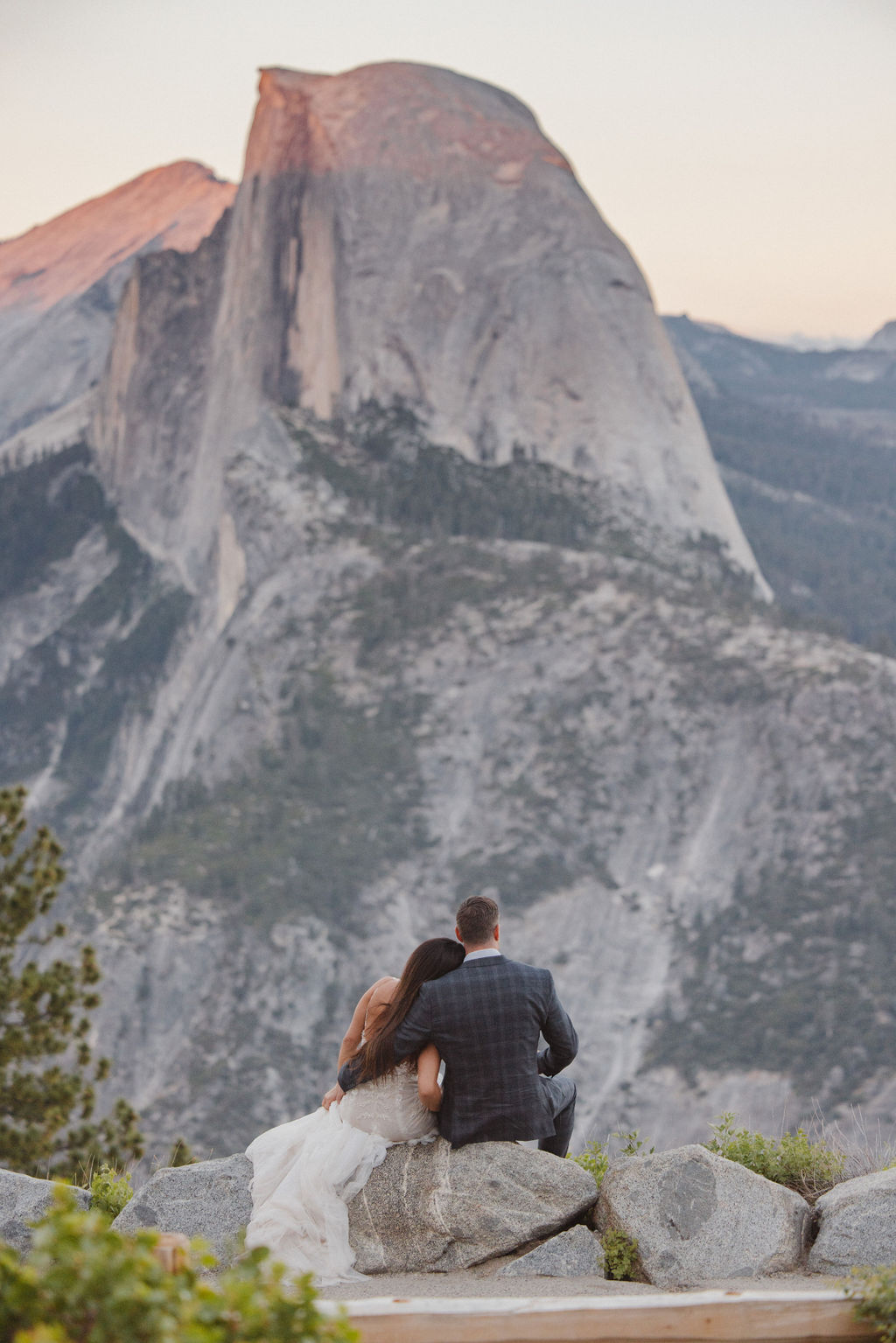 A couple sits on a rock ledge, arm in arm, with their backs to the camera, facing a large, prominent mountain peak in the distance at sunset.