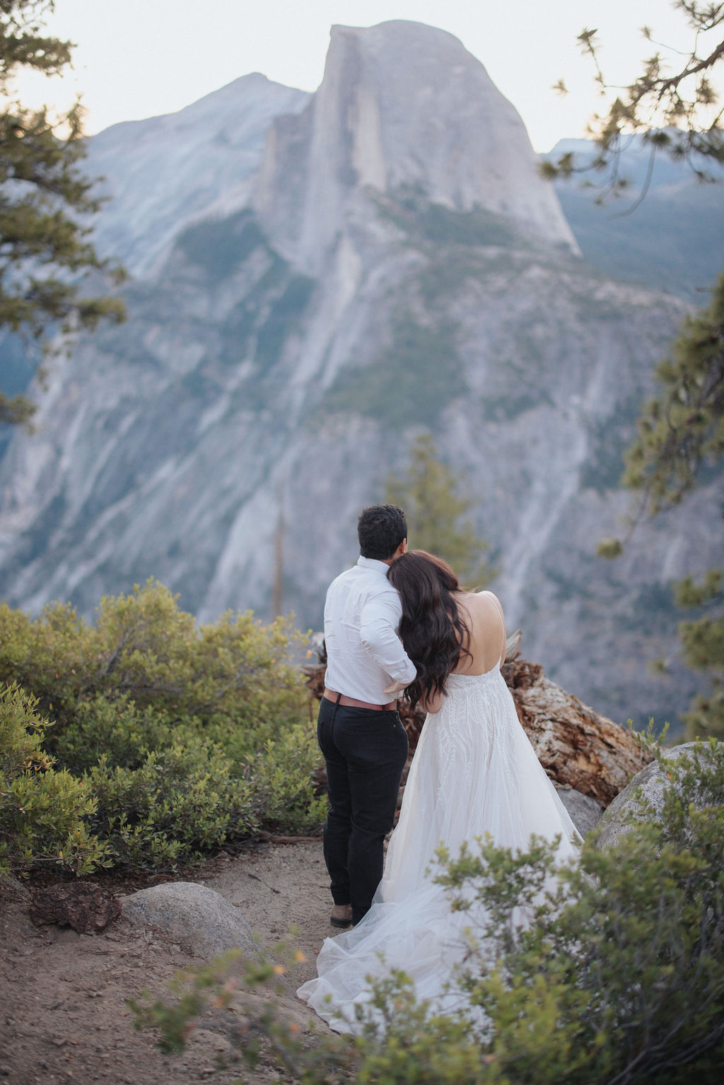 A couple stands at a scenic overlook, facing a mountainous landscape with lush greenery and a prominent rock formation in the background for their Yosemite elopement photos