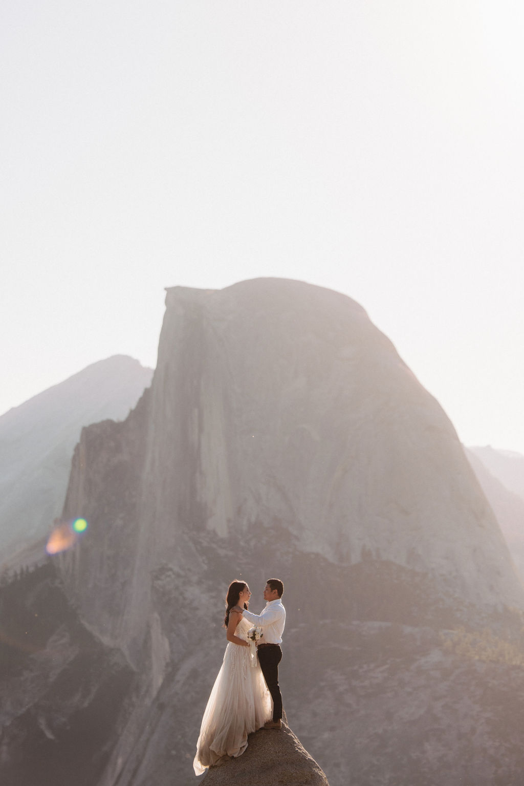 A couple in wedding attire embraces on a rocky ledge, with a large mountain peak in the background under soft, warm sunlight for their Yosemite elopement photos