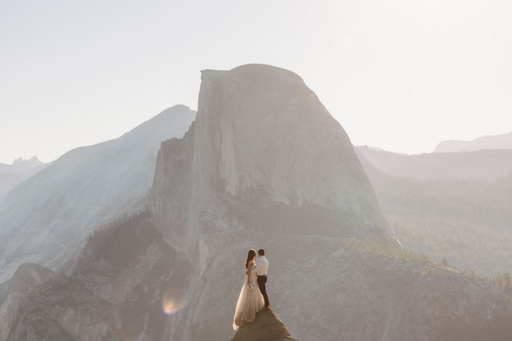 A couple stands on a rocky ledge during sunset; the man, wearing a white shirt and dark pants, holds the hand of the woman, who is in a flowing white dress. A mountain peak is visible in the background for their yosemite elopement photos