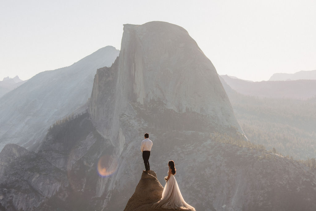 A couple stands on a rocky ledge during sunset; the man, wearing a white shirt and dark pants, holds the hand of the woman, who is in a flowing white dress. A mountain peak is visible in the background for their yosemite elopement photos