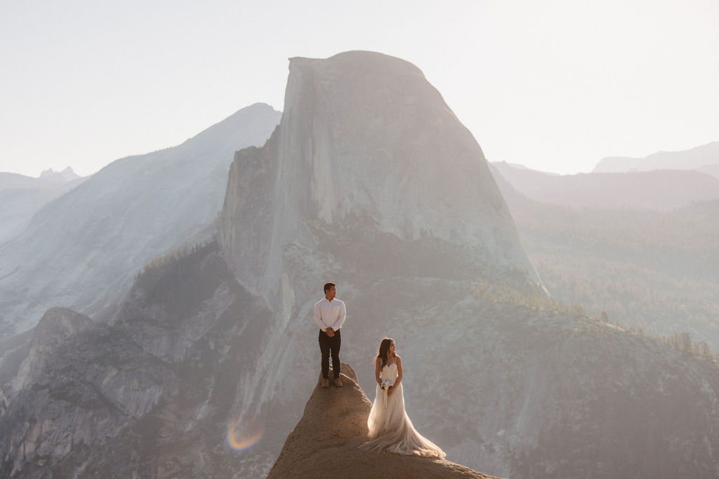 A couple dressed in formal attire stands at the edge of a cliff with a large mountain in the background, under a clear sky.