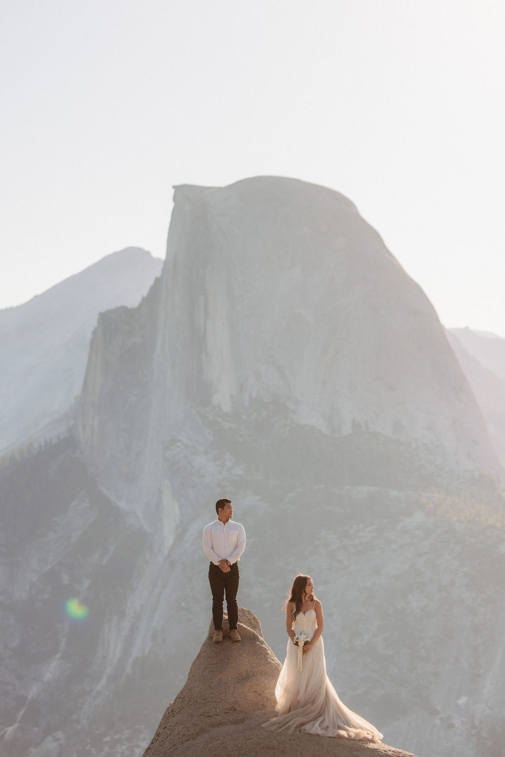 A couple in wedding attire embraces on a rocky ledge, with a large mountain peak in the background under soft, warm sunlight for their Yosemite elopement photos