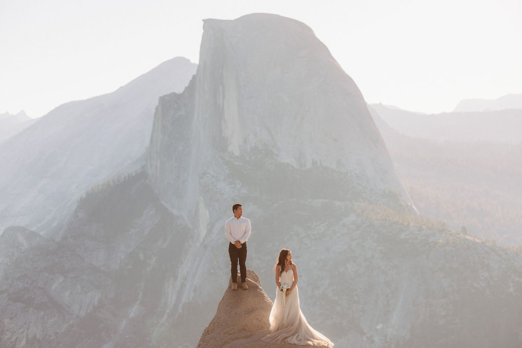 A couple stands on a rocky ledge during sunset; the man, wearing a white shirt and dark pants, holds the hand of the woman, who is in a flowing white dress. A mountain peak is visible in the background for their yosemite elopement photos