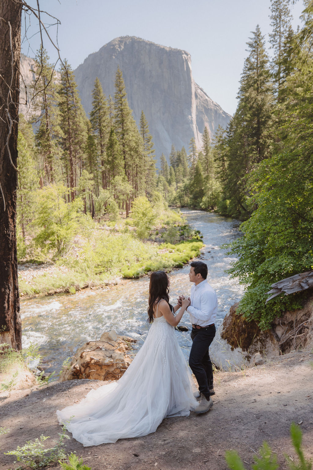 A couple in wedding attire faces each other by a river, surrounded by trees and mountains in the background for their Yosemite elopement photos