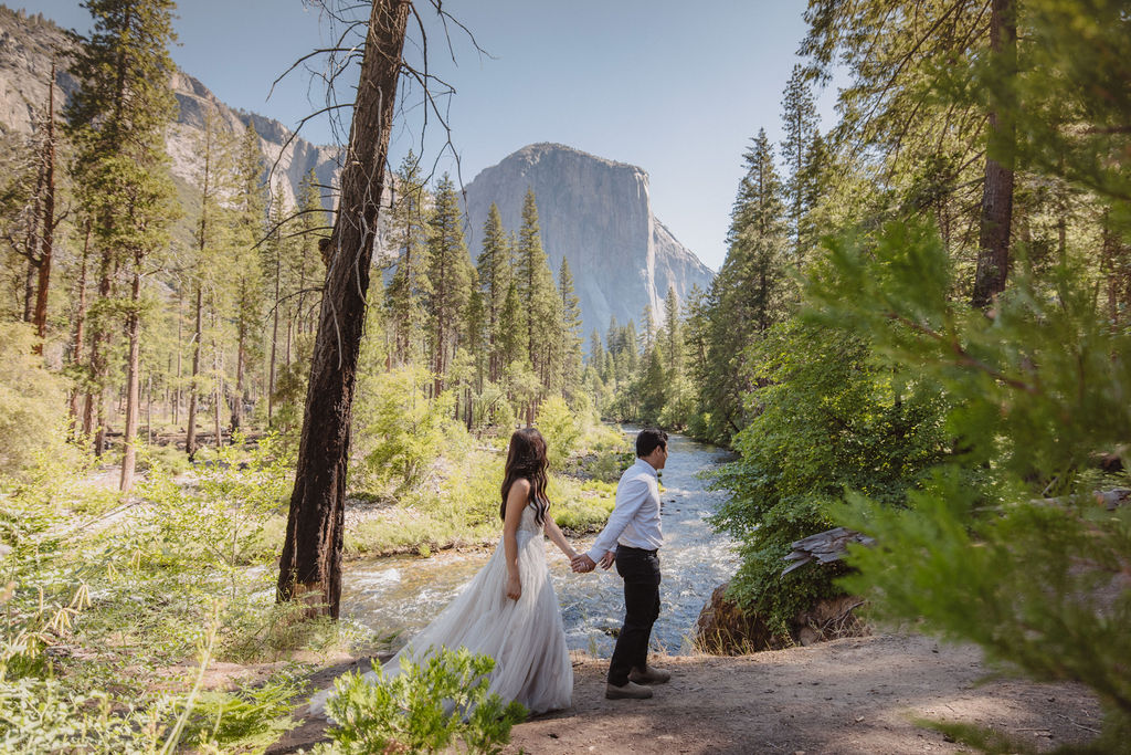 A couple in wedding attire faces each other by a river, surrounded by trees and mountains in the background for their Yosemite elopement photos