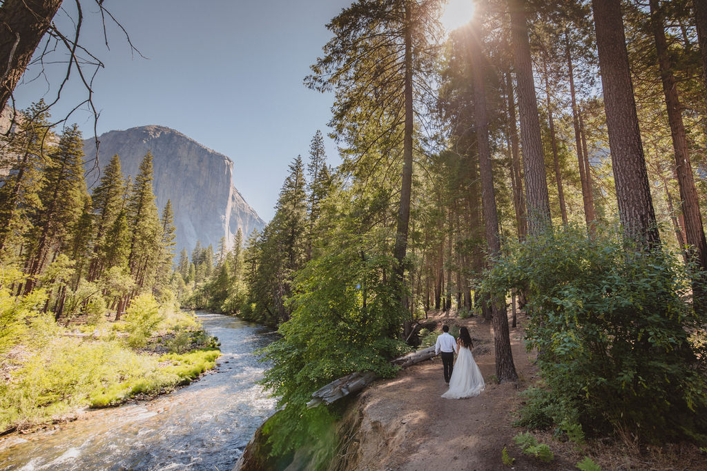 A couple in wedding attire faces each other by a river, surrounded by trees and mountains in the background for their Yosemite elopement photos