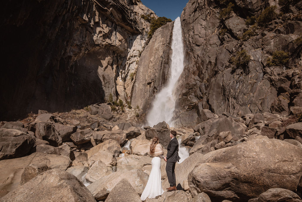 A couple stands on rocks in formal attire with a cascading waterfall behind them at Yosemite falls for their yosemite elopement