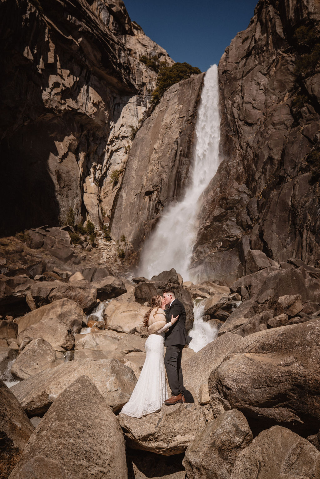 A couple stands on rocks in formal attire with a cascading waterfall behind them at Yosemite falls for their yosemite elopement