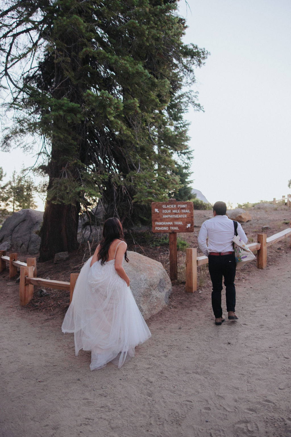 A bride in a white dress and a person in a white shirt and dark pants walk along a dirt path with a large tree and wooden sign in the background for their Yosemite elopement photos