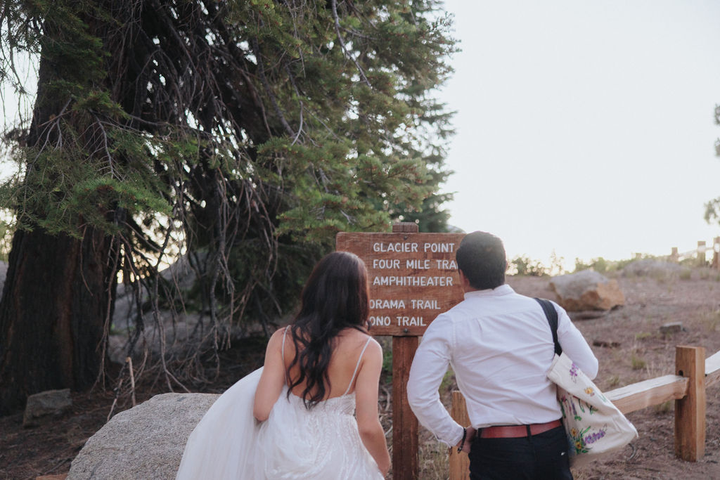 A bride in a white dress and a person in a white shirt and dark pants walk along a dirt path with a large tree and wooden sign in the background for their Yosemite elopement photos