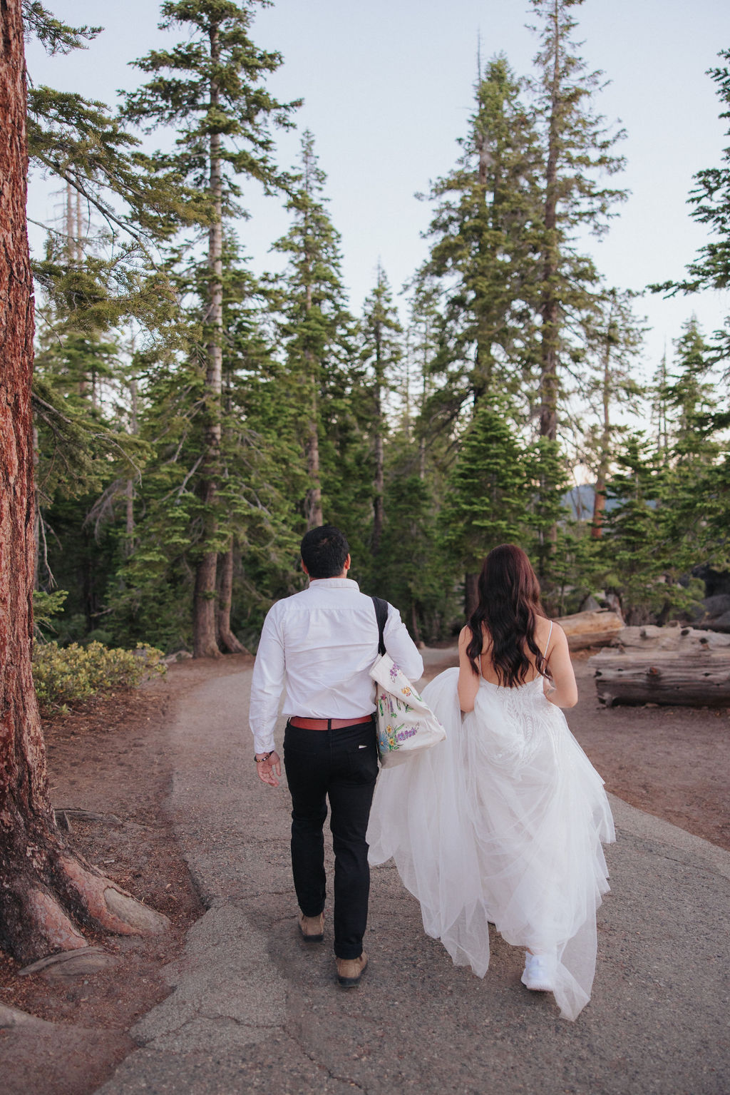 A bride in a white dress and a person in a white shirt and dark pants walk along a dirt path with a large tree and wooden sign in the background for their Yosemite elopement photos