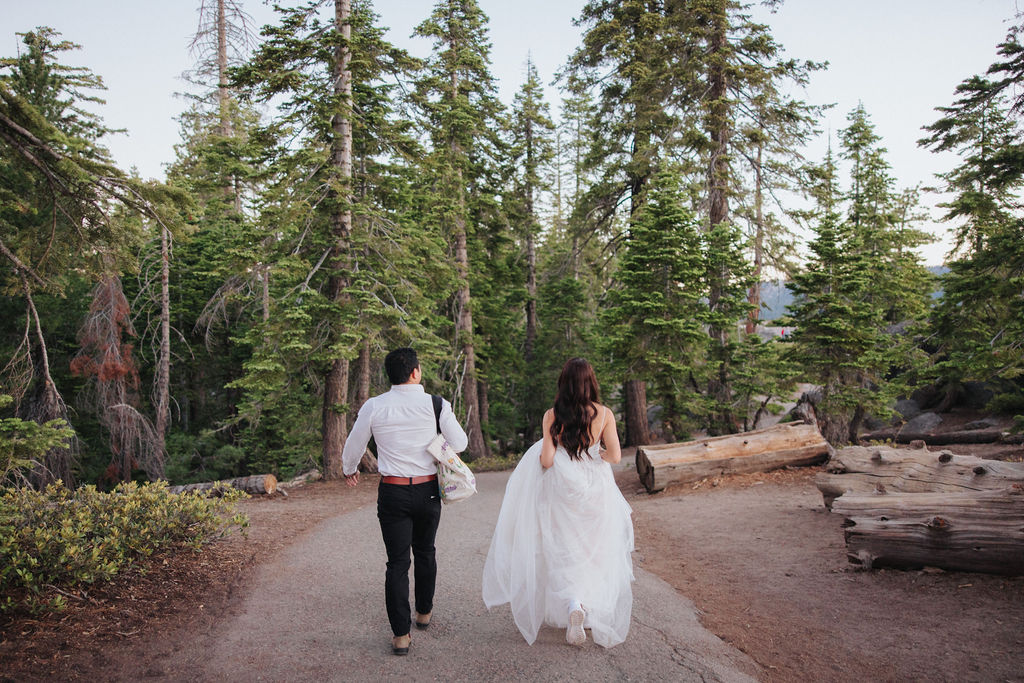 A bride in a white dress and a person in a white shirt and dark pants walk along a dirt path with a large tree and wooden sign in the background for their Yosemite elopement photos