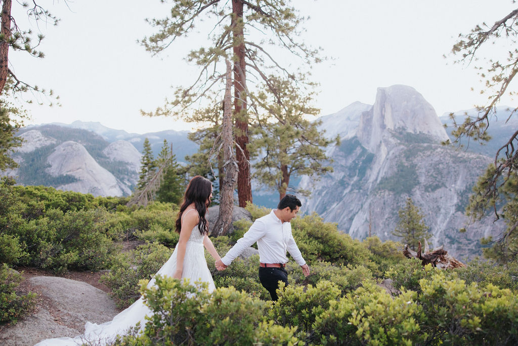 A couple stands at a scenic overlook, facing a mountainous landscape with lush greenery and a prominent rock formation in the background for their Yosemite elopement photos