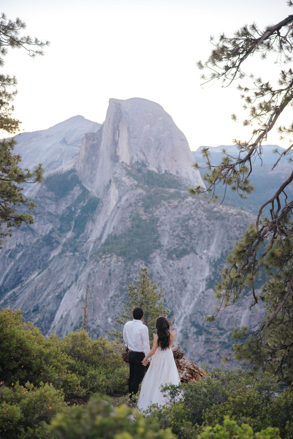 A couple stands at a scenic overlook, facing a mountainous landscape with lush greenery and a prominent rock formation in the background for their Yosemite elopement photos