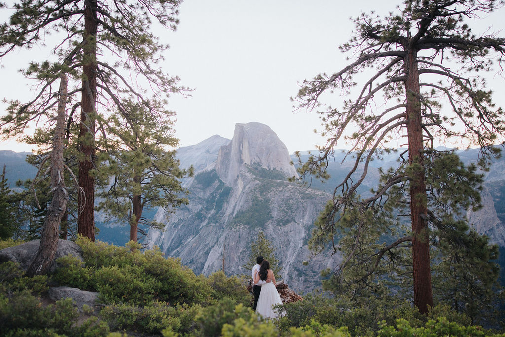 A couple stands at a scenic overlook, facing a mountainous landscape with lush greenery and a prominent rock formation in the background for their Yosemite elopement photos