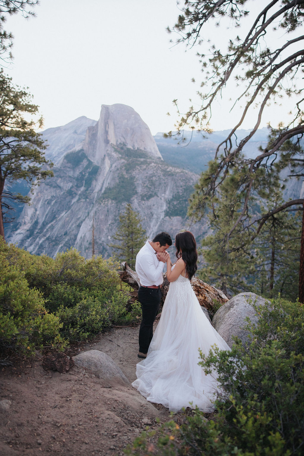 A couple stands at a scenic overlook, facing a mountainous landscape with lush greenery and a prominent rock formation in the background for their Yosemite elopement photos