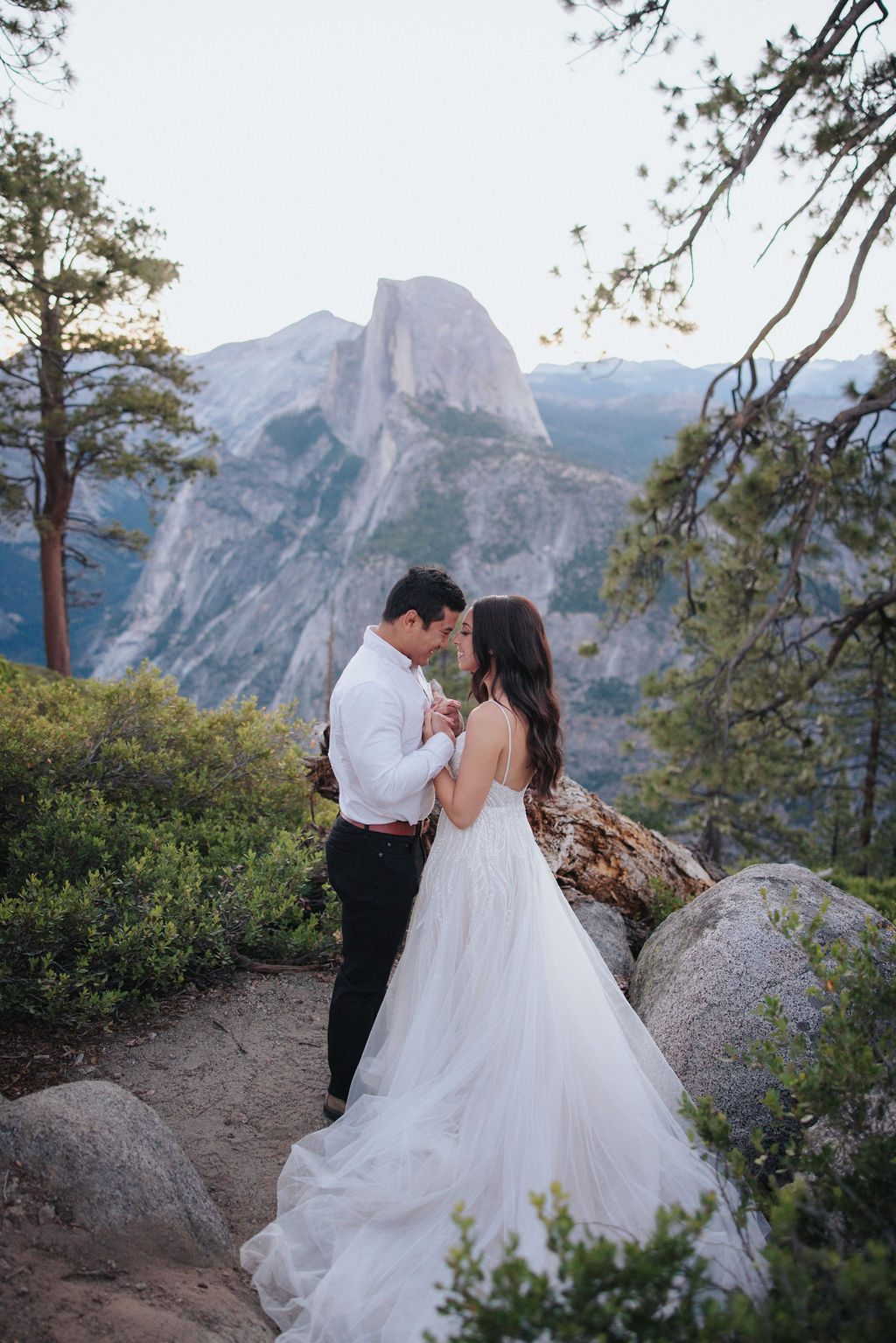 A couple stands at a scenic overlook, facing a mountainous landscape with lush greenery and a prominent rock formation in the background for their Yosemite elopement photos