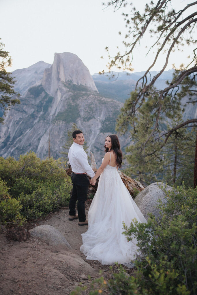 A couple stands at a scenic overlook, facing a mountainous landscape with lush greenery and a prominent rock formation in the background for their Yosemite elopement photos