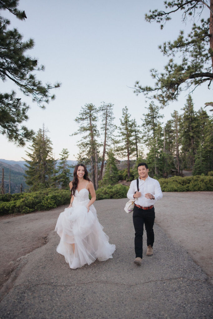 A couple stands at a scenic overlook, facing a mountainous landscape with lush greenery and a prominent rock formation in the background for their Yosemite elopement photos