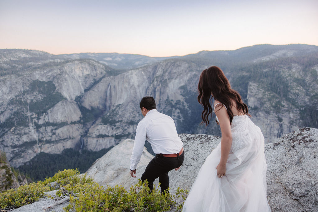 A couple stands at a scenic overlook, facing a mountainous landscape with lush greenery and a prominent rock formation in the background for their Yosemite elopement photos