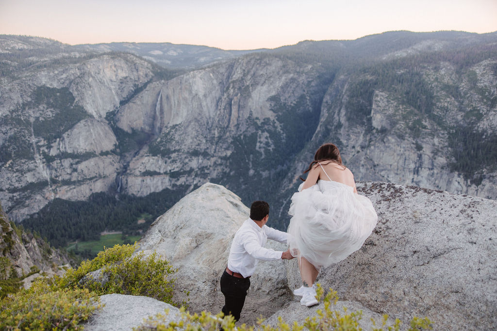 A couple stands at a scenic overlook, facing a mountainous landscape with lush greenery and a prominent rock formation in the background for their Yosemite elopement photos