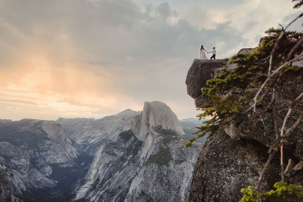 A couple stands on the edge of a cliff with mountainous terrain and a dramatic sky in the background at Glacier point for their elopement at yosemite