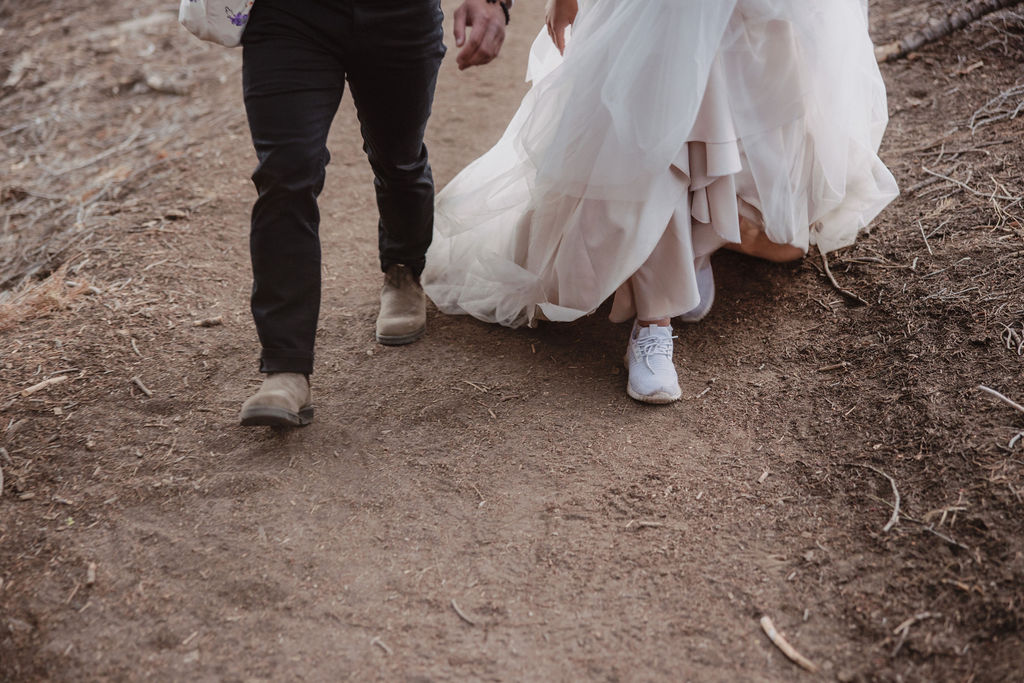 A couple, seen from behind, walks hand in hand on a road through a forest. The woman is wearing a white dress, and the man is in a white shirt and dark pants.