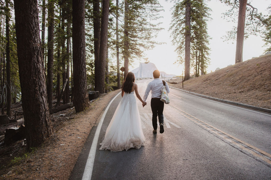 A couple, seen from behind, walks hand in hand on a road through a forest. The woman is wearing a white dress, and the man is in a white shirt and dark pants.