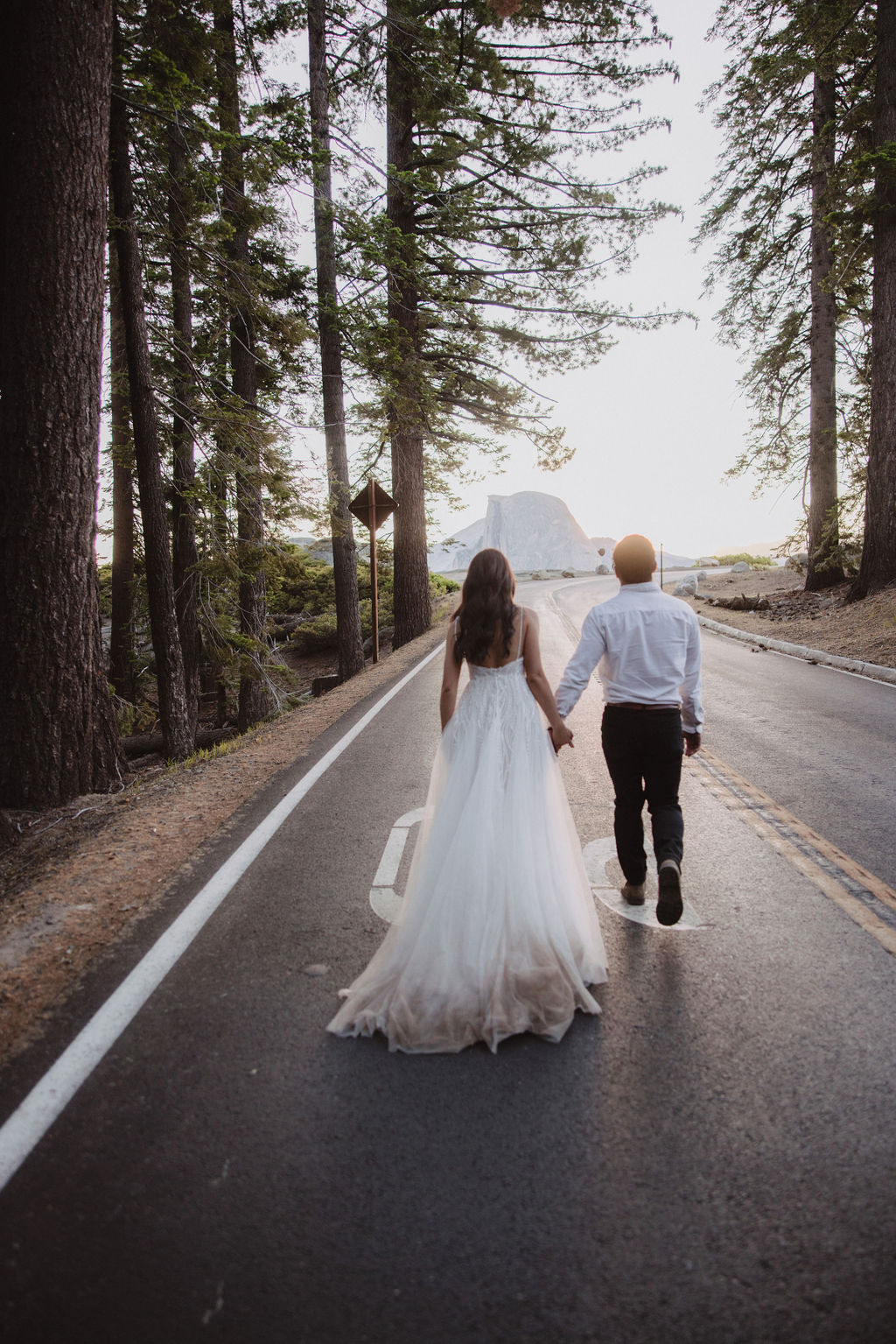 A couple, seen from behind, walks hand in hand on a road through a forest. The woman is wearing a white dress, and the man is in a white shirt and dark pants.